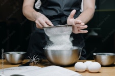 Cropped image of female pastry chef sifting flour for homemade baking, woman in apron holding sieve surrounded by baking... 