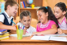 Three Primary School Children Working Together with Toy Construction Blocks in a Classroom, the Girl Reading Instructions from a... 