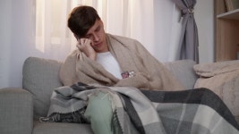 온라인 약국. 아픈 사람. 모바일 상담. 기분 나쁜 남자는 소파 조명 홈 인테리어에 앉아 손에 태블릿을 들고 격자 무늬 말하는 스마트폰에... 