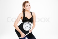 Happy fitness woman holding weighing machine isolated on a white background and looking at camera | Stock Photo | Colourbox