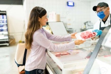 Buying The Best Fresh Steak At The Market. Happy Young Woman At The Supermarket Asking A Salesman For Raw Meat To Cook For Dinner...